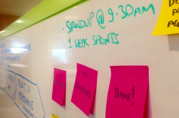 Picture of a whiteboard which features the text "Standup @ 9.30am, 1 week sprints". The board also has postit notes attached to it that say "Planned", "In progress", and "Done!"