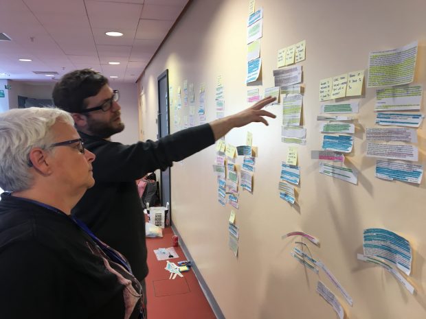 Two colleagues discussing various flowcharts on a wall.